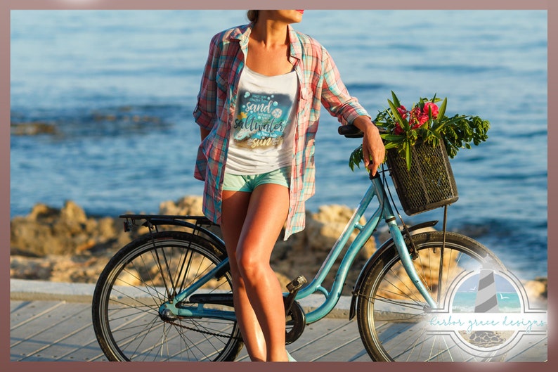 May include: A woman in a white tank top with the text "sand, saltwater, sun" and a blue and white graphic design stands next to a blue bicycle with a basket of flowers. The bicycle is parked on a wooden boardwalk near the ocean.