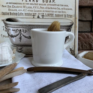 May include: A white ceramic shaving mug with a brown shaving brush inside. The mug is sitting on a white cloth with a vintage advertisement for Pears' Soap in the background. There is a straight razor and wooden sticks on the cloth.