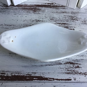May include: White ceramic serving dish with a slightly curved shape. The dish has a smooth, glossy finish and two raised decorative elements on either side.