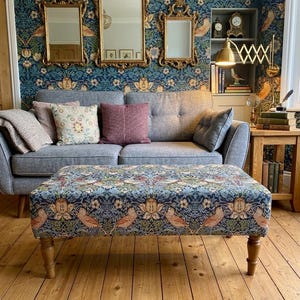 May include: A rectangular ottoman featuring a blue, green, and orange floral and bird tapestry fabric upholstery with four turned wooden legs. The ottoman is positioned in front of a grey sofa with various throw pillows and a patterned wallpaper.