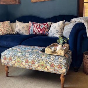 May include: A rectangular ottoman upholstered in a multi-coloured fabric featuring a dense pattern of birds, flowers, and foliage in shades of blue, yellow, and red, supported by four turned wooden legs, with a woven tray holding a small plant.