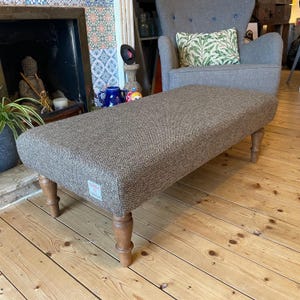 May include: A rectangular ottoman upholstered in a brown and black herringbone patterned fabric. The piece features four turned wooden legs and a small rectangular fabric label attached to the lower front corner of the upholstered frame.