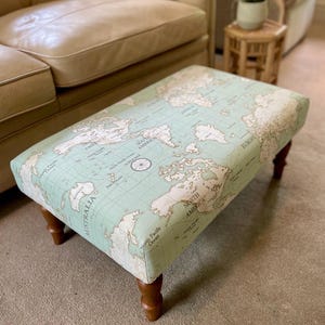 May include: A rectangular ottoman upholstered with a world map design. The map is in beige and brown on a light green background. The ottoman has four dark wooden legs and is placed in a living room setting.