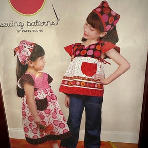 May include: A sewing pattern package featuring two young girls modeling a tunic-style dress and shirt with headscarves. The package includes the text "modkid frida" and "sewing patterns by Patty Young". Sizes 2T-7 are indicated.