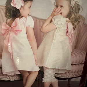 May include: Two young girls in matching dresses. One girl is wearing a light pink dress with a large pink bow on the back and a pink flower headband. The other girl is wearing a similar dress with a white lace bow and white polka dot pants.