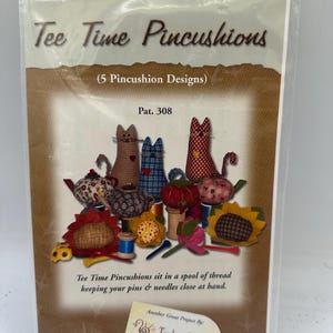 May include: A sewing pattern package titled Tee Time Pincushions featuring five pincushion designs. The cover displays fabric cats, teapots, and sunflowers sitting on thread spools. The text reads Tee Time Pincushions, 5 Pincushion Designs, Pat. 308.