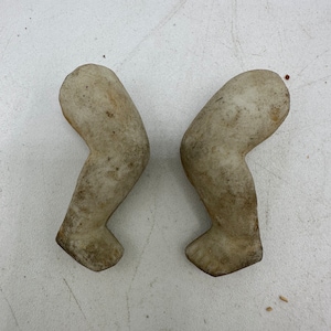 May include: Two ceramic doll arms, painted in a light gray color, are shown against a white background. The arms are bent at the elbow and the hands are closed.