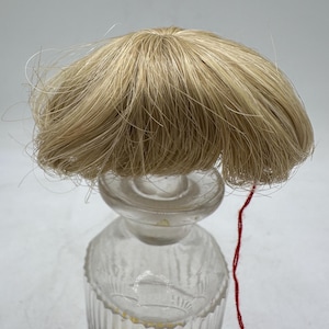 May include: A clear glass perfume bottle with a decorative design and a blonde hairpiece attached to the stopper. A red string is attached to the hairpiece. The bottle has a ribbed design and a round stopper.