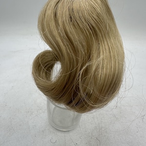 May include: A light blonde wig with a curled section. The wig is styled with a side part and has a natural-looking hairline. The hair is a warm, light blonde color and appears to be made of synthetic fibers.