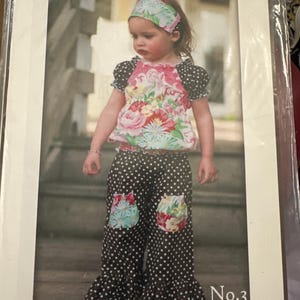 May include: A child's outfit featuring a floral top, polka dot pants, and a matching headband. The top has a pink floral pattern with black polka dot sleeves. The pants are black with white polka dots and floral patches. The text "the OLIVIA top" is visible.