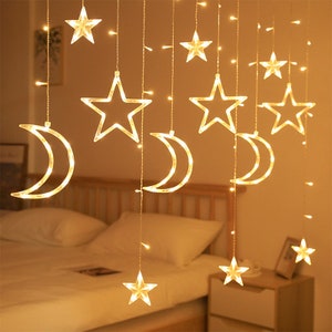 May include: A string of warm white fairy lights with star and crescent moon shapes. The lights are hanging from a ceiling and are illuminating a bedroom.