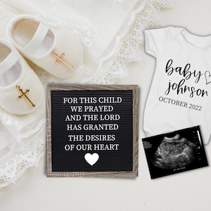 Op de afbeelding: Een witte romper met de tekst "baby johnson OCTOBER 2022" en een klein hartje. Een zwart-witte echografie, een wit letterbord met de tekst "FOR THIS CHILD WE PRAYED AND THE LORD HAS GRANTED THE DESIRES OF OUR HEART", een paar witte babyschoentjes met gouden kruisen en een wit en gouden kralenarmband met een kruisbedel.