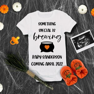 May include: White onesie with the text "Something special is brewing" and a black cauldron with an orange heart. Below the cauldron is the text "Baby Sanderson coming April 2022". The onesie is surrounded by orange and white pumpkins, a pacifier, a broom, and a sonogram image.