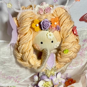 Op de afbeelding: Een handgemaakte pop met garen haar, versierd met een bloemenkroon en decoratieve geweien. De pop heeft een stoffen gezicht en is gekleed in een lavendel en witte outfit, met een vlinder accent. Het ontwerp is grillig en sprookjesachtig.