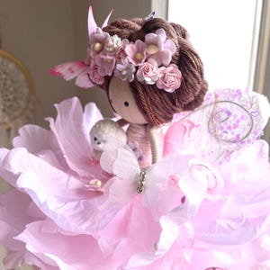 Könnte beinhalten: Eine handgefertigte Feenpuppe mit einem Holzkopf, braunem Garnhaar und einem rosa Blumenkranz. Die Puppe trägt ein rosa Kleid aus geschichteten Blütenblättern und hat Schmetterlingsflügel. Ein kleines Stofftier wird von der Puppe gehalten.