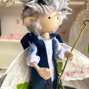 May include: A handcrafted fairy doll with a wooden head, white hair, and blue accents. The doll wears a navy blue vest over a white shirt, with delicate wings and a gold-colored staff. The fairy has a whimsical design with intricate details.