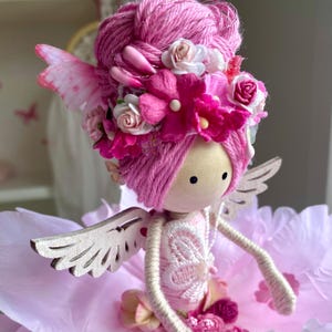 May include: A handcrafted fairy doll with a wooden head, pink yarn hair, and delicate wooden wings. The doll is adorned with a floral crown of pink and white flowers. The fairy is dressed in a pink dress and sits on a pink surface.