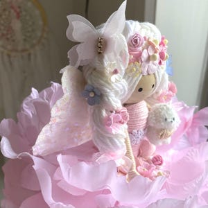 May include: A handmade doll with white yarn hair, adorned with pink and blue flowers, and a butterfly hair accessory. The doll is dressed in a pink outfit and holds a small white dog. It is set on a bed of pink petals.