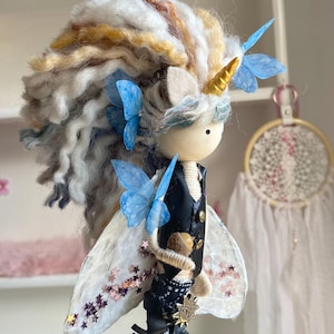 May include: A handcrafted wooden doll with a unicorn horn and butterfly wings. The doll features a multi-coloured yarn mane, blue butterfly accents, and a black waistcoat. It holds a small acorn and has a gold horn. The wings are white with pink star details.