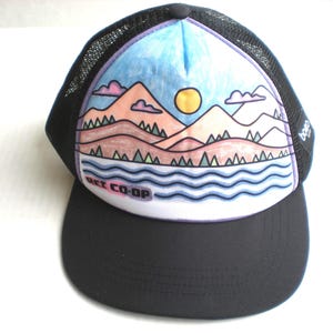 May include: Black trucker hat with a mesh back and a curved brim. The front panel features a colorful illustration of a mountain landscape with a sun, water, and the text "REI CO-OP". The hat has a black brim and a white front panel.