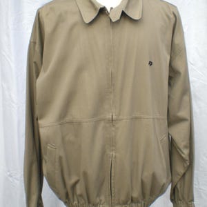 May include: A tan jacket with a collared neckline and a zipper closure. The jacket has long sleeves, a small logo on the chest, and two front pockets. The bottom hem is elasticized.