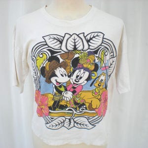 May include: Off-white t-shirt featuring a colorful graphic of Mickey and Minnie Mouse embracing, with Pluto nearby. The design includes tropical flowers and foliage, framed by a decorative border. The shirt has short sleeves.
