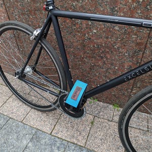 Könnte beinhalten: Ein mattschwarzer Fahrradrahmen mit dem Wort EXCELSIOR auf dem Unterrohr und DANDY auf dem Oberrohr. Ein hellblauer Pedalgurt mit einem braunen Lederaufnäher mit der Aufschrift Black Box ist am schwarzen Fahrradpedal befestigt.