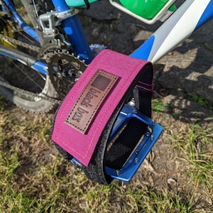Op de afbeelding: Close-up van een fietspedaal met een magenta voetband. De band heeft een zwarte achterkant en een bruin leren label met de tekst "black box". Het pedaal is blauw en bevestigd aan een blauw-wit fietsframe.