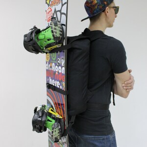 May include: A person wearing a black t-shirt and blue jeans is carrying a snowboard with colourful stickers and bindings. The snowboard has the text "Lilo" on it.