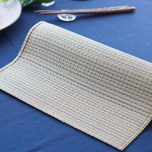 May include: A rolled up placemat with a woven texture in beige and blue. The placemat is on a blue surface.