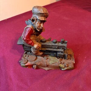 May include: A detailed figurine of a man in a cap and red vest, working on a miniature train engine. The figurine has a weathered appearance with brown, green, and red tones, and sits on a detailed base.