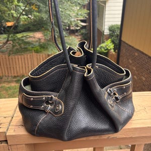 May include: A black pebbled leather handbag with two long shoulder straps and tan contrast stitching. The bag features a gathered, open-top design with silver-toned metal hardware, including side buckles and circular accents on the exterior panels.