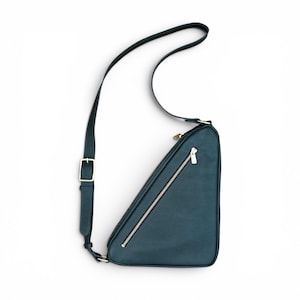 May include: A dark teal triangular crossbody bag with a long adjustable shoulder strap featuring a gold-toned buckle. The bag has a diagonal zippered pocket on the front with a metallic zipper pull and a smooth, matte finish exterior surface.