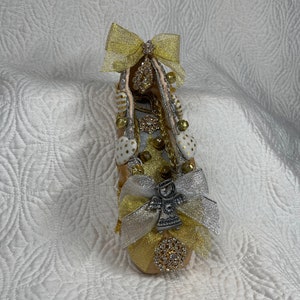 Pode incluir: Uma sapatilha de balé decorada em dourado e branco com um laço, sinos e um amuleto de anjo.