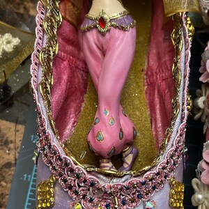 Könnte beinhalten: Ein lilafarbener Ballettschuh, verziert mit goldenen Verzierungen, Strasssteinen und einem goldenen Anhänger mit baumelnden Münzen. Im Schuh befindet sich eine Miniaturfigur einer Frau in einem rosa Kostüm.