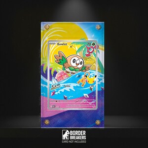 May include: A framed Rowlet Pokémon card with a colorful tropical beach scene background. The card features a cartoon owl with green and brown feathers. The background includes palm trees, waves, and a yellow sky. The card is in a clear acrylic case.