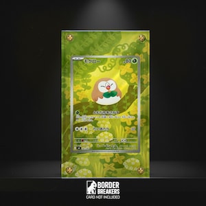 May include: A framed Pokémon trading card featuring Rowlet, a brown and white owl-like creature with a green bow tie. The card is set against a green background with floral and leaf patterns, and the frame is secured with gold-colored screws.