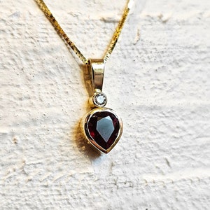 Op de afbeelding: Een gouden ketting met een hartvormige hanger met een rode edelsteen en een klein, helder diamant accent.