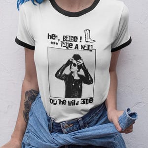 May include: White ringer t-shirt with black trim featuring a black and white graphic. The graphic includes text and an illustration of a person wearing sunglasses. The text reads "Hey, babe! ... Take a walk on the wild side."