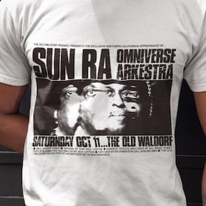 Puede incluir: Camiseta blanca con un gráfico en blanco y negro de Sun Ra y la Omniverse Arkestra. El diseño incluye texto que anuncia una actuación en The Old Waldorf el sábado 11 de octubre.