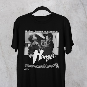 Op de afbeelding: Zwart t-shirt met een zwart-wit afbeelding uit de film "The Hunger". De afbeelding bevat de titel van de film, namen van de cast en de zin "Nothing human loves forever."