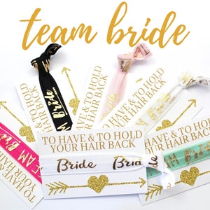May include: A set of six hair ties with gold glitter and the text "To Have & To Hold Your Hair Back" and "Team Bride". The hair ties are on white cards with gold glitter hearts and arrows.