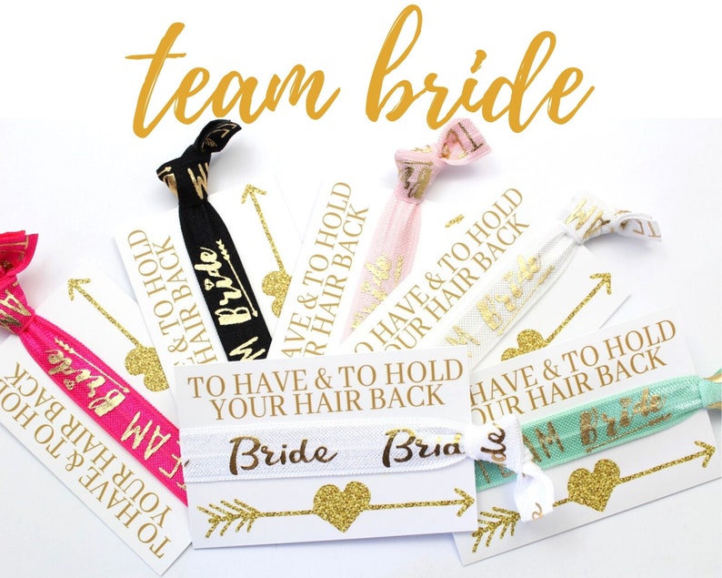 May include: A set of six hair ties with gold glitter and the text "To Have & To Hold Your Hair Back" and "Team Bride". The hair ties are on white cards with gold glitter hearts and arrows.