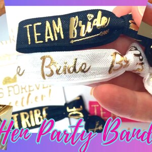May include: A set of elastic hair ties with gold lettering. The hair ties are black, white, and gold. The text on the hair ties includes "Team Bride", "Bride Tribe", and "Friends Forever Together".