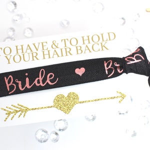May include: A black elastic hair tie with the word "Bride" in pink glitter lettering and a small pink heart. The hair tie is on a white card with the text "To Have & To Hold Your Hair Back" in gold lettering and a gold glitter arrow with a gold glitter heart.