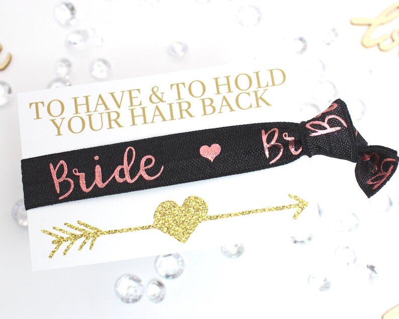 May include: A black elastic hair tie with the word "Bride" in pink glitter lettering and a small pink heart. The hair tie is on a white card with the text "To Have & To Hold Your Hair Back" in gold lettering and a gold glitter arrow with a gold glitter heart.