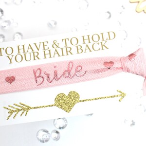 May include: A pink hair tie with gold glitter lettering that says "Bride" and the text "To Have & To Hold Your Hair Back".