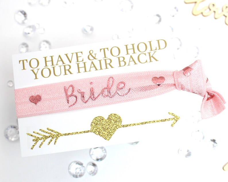 May include: A pink hair tie with gold glitter lettering that says "Bride" and the text "To Have & To Hold Your Hair Back".