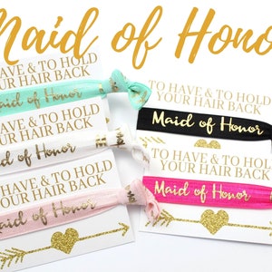 May include: Set of five hair ties with the text "Maid of Honor" and the phrase "To Have & To Hold Your Hair Back" printed on them. The hair ties are in various colors, including pink, black, white, and green.