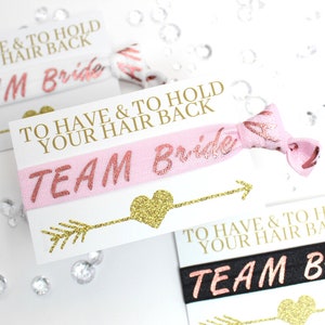 May include: Pink and black hair ties with gold glitter accents. The hair ties have the text "Team Bride" and "To Have & To Hold Your Hair Back" printed on them.